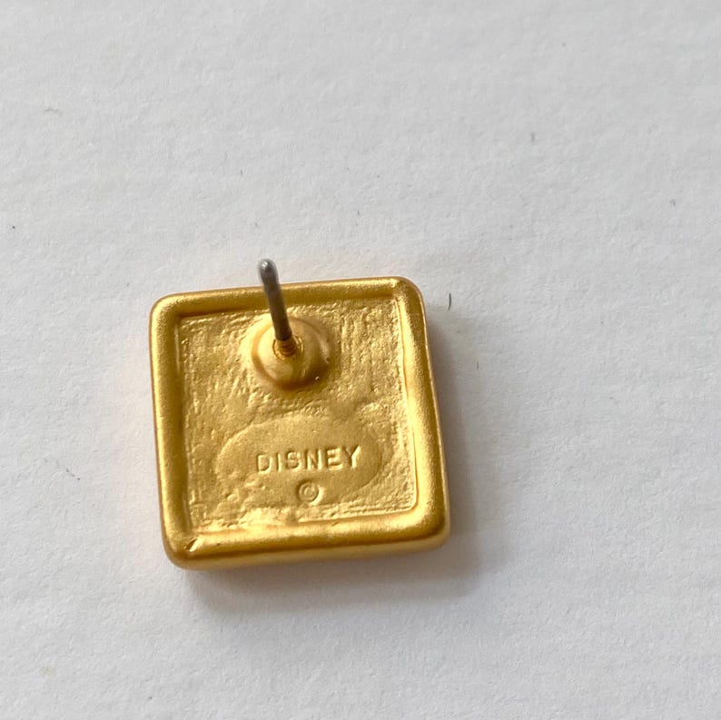May include: Gold-toned square earring with a post back. The back of the earring has the word "DISNEY" engraved on it.