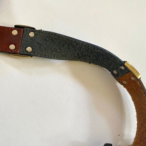 May include: A brown leather belt with a brass buckle and rivets. The belt is partially unbuckled and the underside is made of dark blue leather.