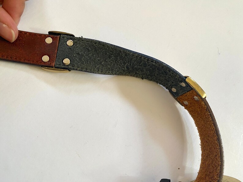 May include: A brown leather belt with a brass buckle and rivets. The belt is partially unbuckled and the underside is made of dark blue leather.