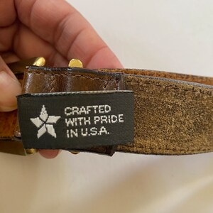 May include: A brown leather belt with a gold buckle and a black label that reads "CRAFTED WITH PRIDE IN U.S.A."