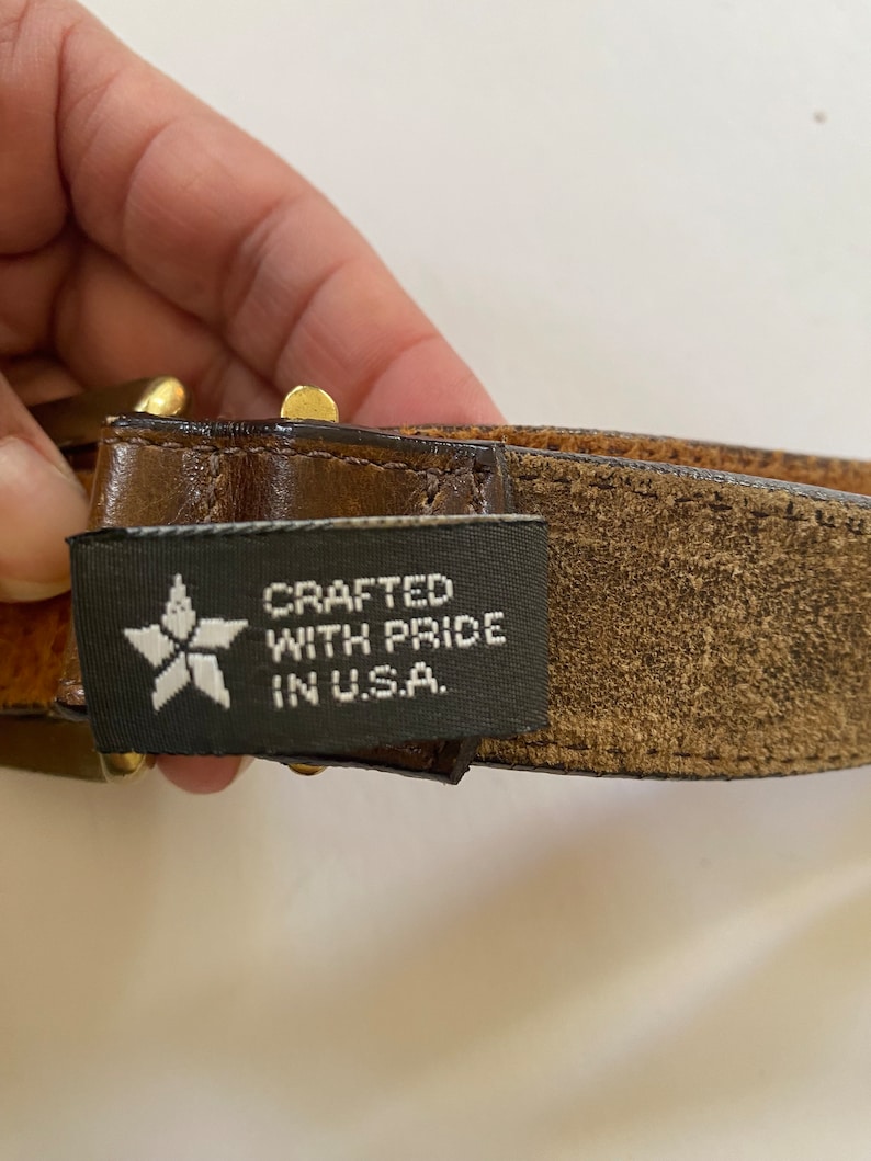 May include: A brown leather belt with a gold buckle and a black label that reads "CRAFTED WITH PRIDE IN U.S.A."