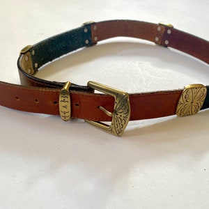 May include: A brown leather belt with a gold metal buckle and decorative accents. The belt has a black leather section with a decorative design.