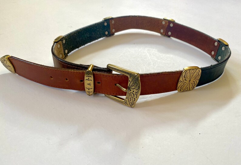 May include: A brown leather belt with a gold metal buckle and decorative accents. The belt has a black leather section with a decorative design.