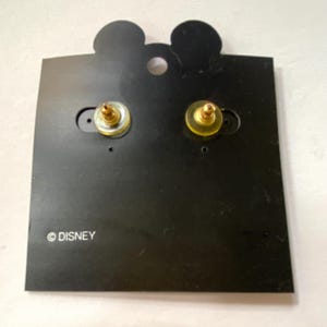 May include: A pair of gold-tone stud earrings with a black background. The earrings are on a black card with the word "DISNEY" printed on it.