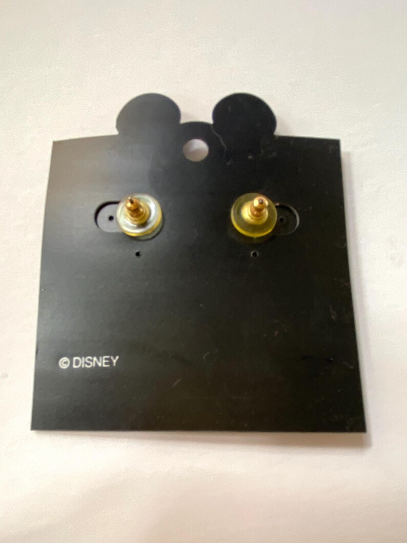 May include: A pair of gold-tone stud earrings with a black background. The earrings are on a black card with the word "DISNEY" printed on it.