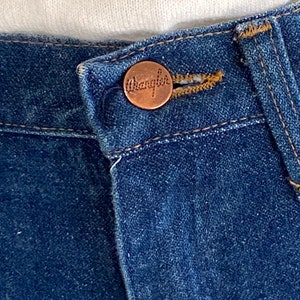 May include: Close-up of a blue denim jean with a copper-colored button that says "Wrangler" on it.