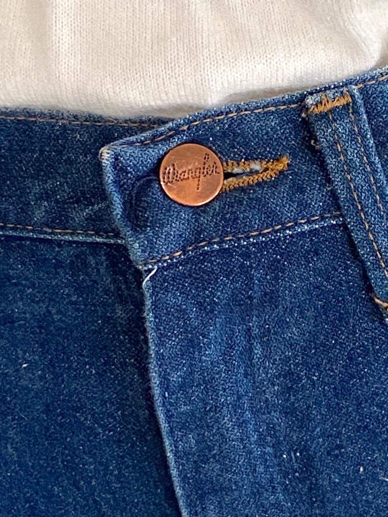 May include: Close-up of a blue denim jean with a copper-colored button that says "Wrangler" on it.
