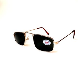 May include: Gold-framed sunglasses with dark lenses. The rectangular lenses are set in a gold frame with a thin, wire-like design. The temples are a dark brown color. A sticker on the lens reads "MAXIMUM UV PROTECTION."