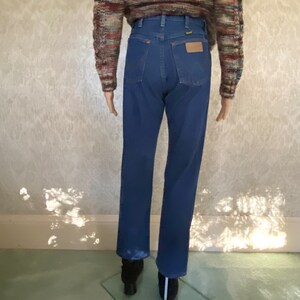 May include: A pair of blue denim jeans with a high waist and a straight leg. The jeans have a brown leather belt loop and a small leather patch on the back pocket.
