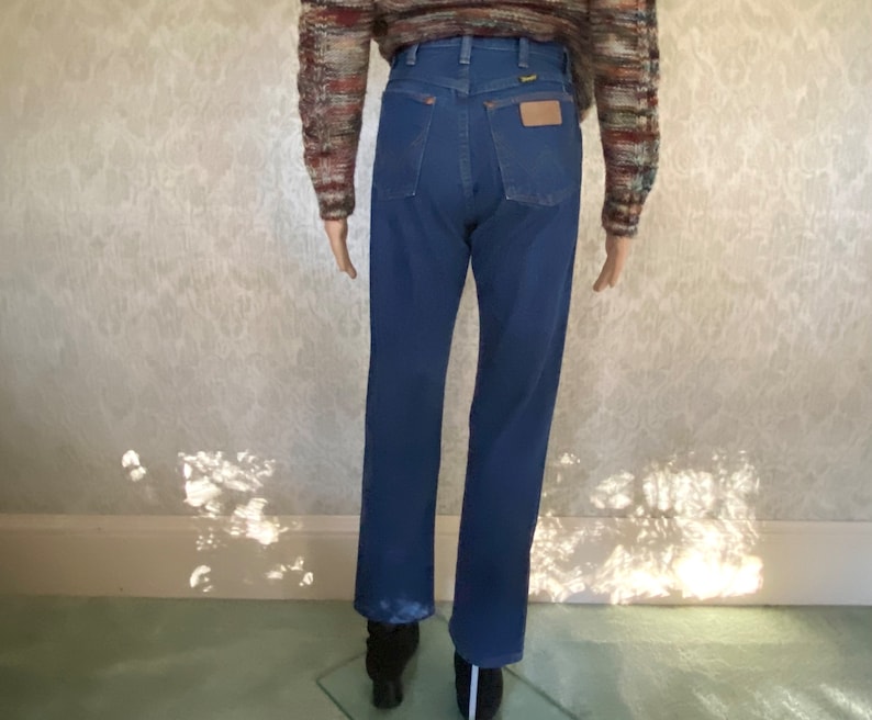 May include: A pair of blue denim jeans with a high waist and a straight leg. The jeans have a brown leather belt loop and a small leather patch on the back pocket.