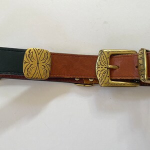 May include: A brown leather belt with two gold-toned buckles and a decorative square buckle. The belt has a black leather section at the beginning.