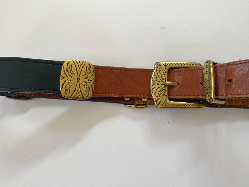 May include: A brown leather belt with two gold-toned buckles and a decorative square buckle. The belt has a black leather section at the beginning.