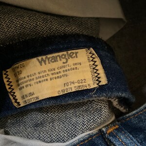 May include: A close-up of a Wrangler brand jeans care label. The label is tan with black text and a black stitched border. The label includes washing instructions, fabric content, and size information. The text on the label reads: "Wrangler, Machine wash with like colors, only chlorine bleach when needed, tumble dry, remove promptly. Made in USA, 100% Cotton, 171WZG, 530, F074-022, 051071 585545."