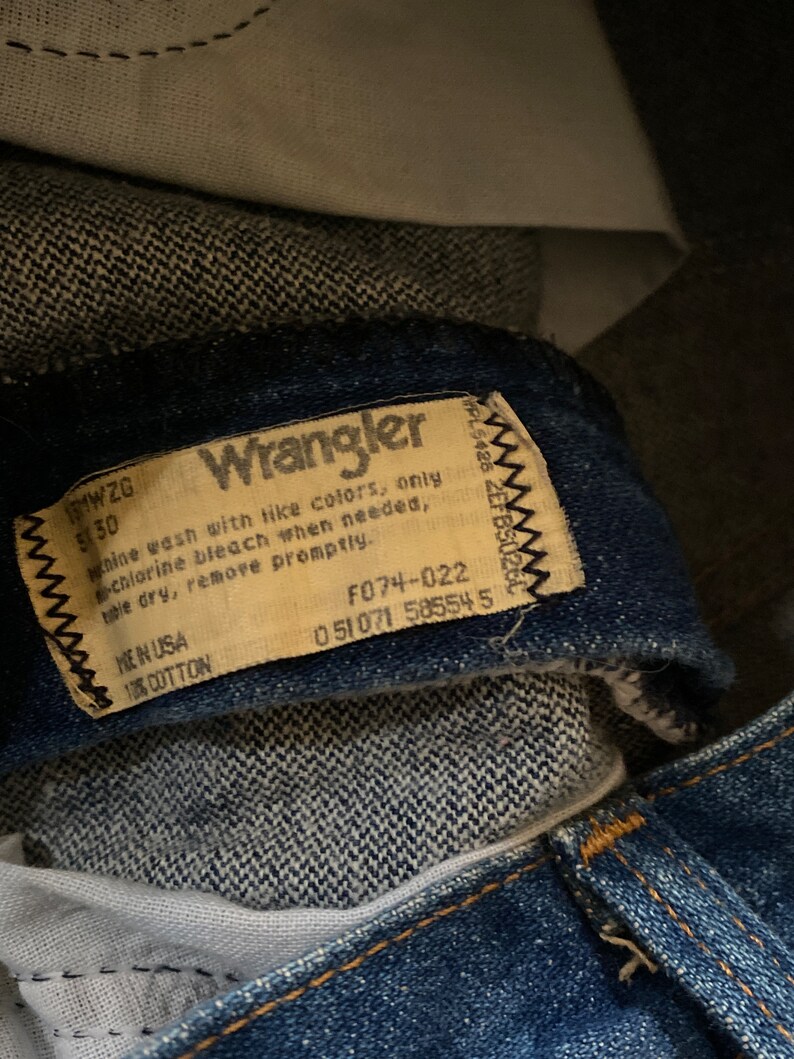 May include: A close-up of a Wrangler brand jeans care label. The label is tan with black text and a black stitched border. The label includes washing instructions, fabric content, and size information. The text on the label reads: "Wrangler, Machine wash with like colors, only chlorine bleach when needed, tumble dry, remove promptly. Made in USA, 100% Cotton, 171WZG, 530, F074-022, 051071 585545."
