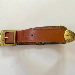 May include: A brown leather belt with a gold metal buckle and tip. The buckle has an ornate design.
