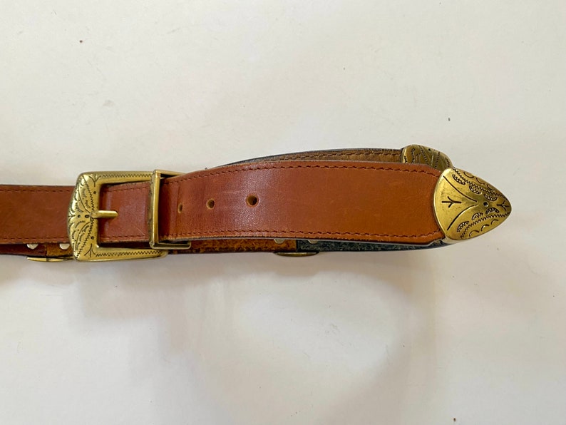May include: A brown leather belt with a gold metal buckle and tip. The buckle has an ornate design.