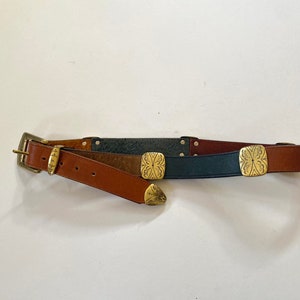 May include: A brown leather belt with a silver buckle and three decorative gold metal accents. The belt is made of three different types of leather: brown, dark brown, and blue.