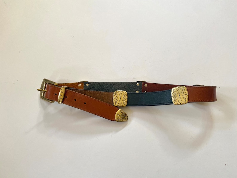 May include: A brown leather belt with a silver buckle and three decorative gold metal accents. The belt is made of three different types of leather: brown, dark brown, and blue.