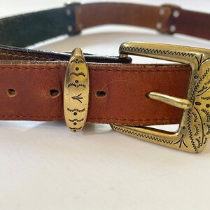 May include: A brown leather belt with a gold metal buckle. The buckle has a decorative design with a geometric pattern.