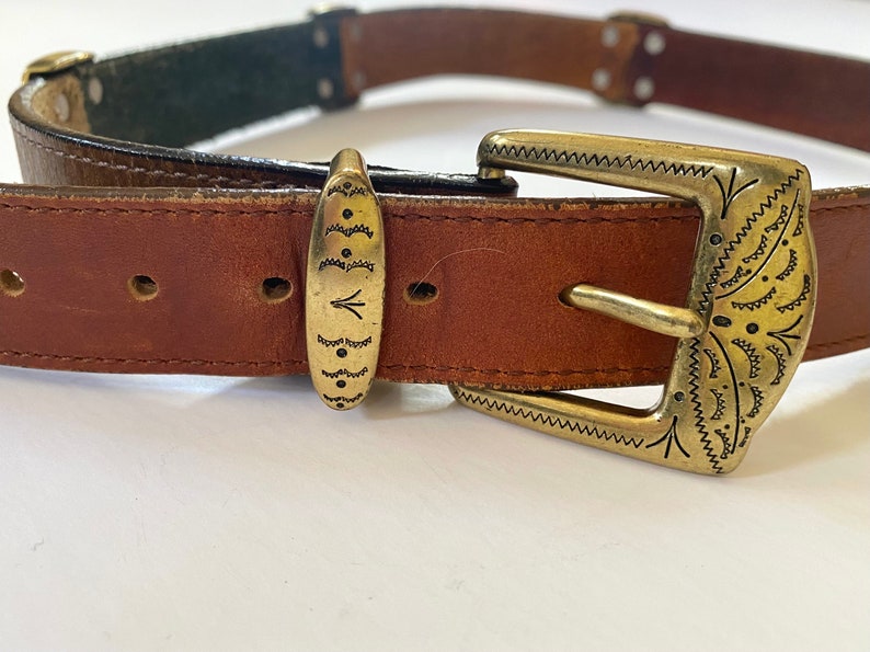 May include: A brown leather belt with a gold metal buckle. The buckle has a decorative design with a geometric pattern.