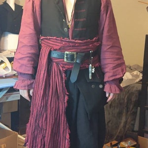 May include: A person dressed as a pirate wearing a black vest, a red shirt, a black belt with a silver buckle, a red striped sash, and black boots. The pirate is wearing a black tricorn hat with a feather and has a long beard.
