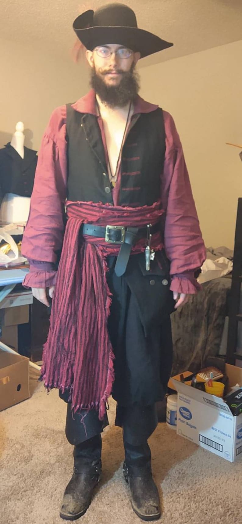 May include: A person dressed as a pirate wearing a black vest, a red shirt, a black belt with a silver buckle, a red striped sash, and black boots. The pirate is wearing a black tricorn hat with a feather and has a long beard.