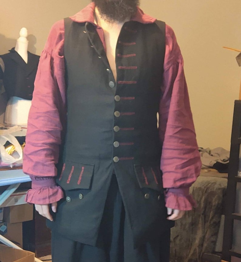 May include: A black vest with red trim and gold buttons. The vest has a pocket on each side and is worn over a pink shirt.