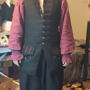 May include: A man wearing a black vest with red trim and matching black pants. He is wearing a burgundy shirt with white cuffs. He has a beard and is wearing glasses.