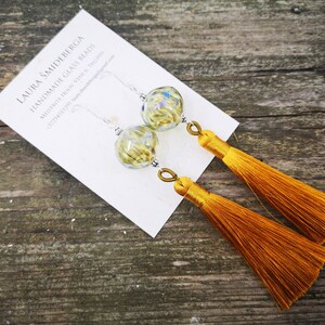 Könnte beinhalten: Ein Paar Ohrringe mit goldenen Quasten und schimmernden grünen Glasperlen. Die Ohrringe befinden sich auf einer weißen Karte mit dem Text "LAURA SMIDBERGA HANDMADE GLASS BEADS MINNEAPOLIS, MINN. SHOP & GALLERY"