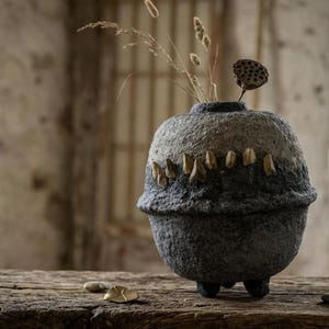 May include: A round, textured, dark grey and charcoal vessel with a wide centre ridge and small feet. The upper section features a row of small, light-coloured, tooth-like protrusions. Dried grasses and a lotus seed pod emerge from the top opening.