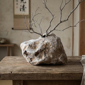 May include: A natural, irregularly shaped stone vase with a rough, textured surface in shades of beige, brown, and white. The stone features a central opening at the top, which holds a bare, branching twig extending upwards from the centre.
