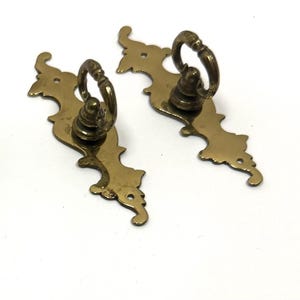 May include: Four brass-toned metal drawer pulls with ornate, elongated backplates and circular drop handles. The set includes a collection of small metal nails and threaded bolts with flat heads, intended for mounting the hardware to furniture.