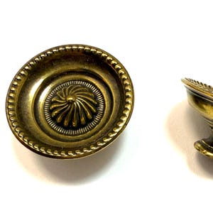 May include: Two antique brass-toned cabinet knobs. One knob is viewed from above, showcasing a central spiral design surrounded by concentric circles and a beaded edge. The other knob is on its side, revealing a pedestal base.