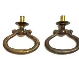 May include: Two brass-toned metal drawer pulls with a drop-ring design and a central mounting post, accompanied by two matching threaded metal screws. The hardware features a weathered, aged finish with visible patina on the circular rings.
