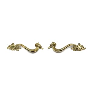 May include: Two ornate, gold-colored drawer pulls with a curved design. Each pull features a central handle with decorative leaf and scroll details. The pulls are set against a plain white background.
