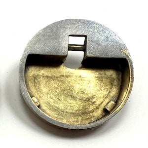May include: A circular metal object with a silver outer ring and a gold-colored interior. The object has a rectangular opening at the top and a curved bottom. The object appears to be a component or part of a larger mechanism.