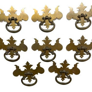 May include: A set of seven brass drawer pulls featuring ornate, symmetrical backplates with decorative leaf-like edges and matching drop handles. The hardware includes a small collection of metal screws for installation, all arranged on a plain surface.