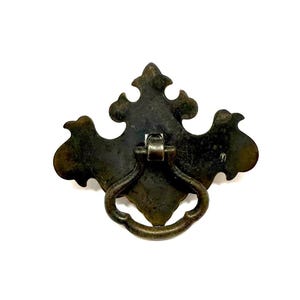 May include: A dark bronze-toned metal drawer pull featuring a decorative backplate with ornate, symmetrical curves and a central drop-style ring handle. The hardware exhibits a weathered, aged patina with visible dark brown and metallic highlights.