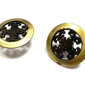 May include: Two antique brass cabinet knobs. Each knob features a circular design with an ornate, dark bronze filigree center. The knobs have a warm, aged patina, suggesting vintage or antique origins. The knobs are set against a white background.
