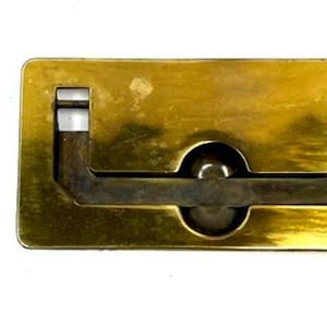 May include: A rectangular brass flush mount drawer pull with a hinged drop handle that rests in a central circular indentation. The hardware features a polished gold-tone finish and includes two small matching metal screws for installation.