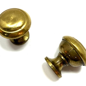 May include: Two antique brass cabinet knobs. The knobs have a rounded, mushroom-like shape with a circular base and a slightly domed top. The brass has a warm, aged patina, suggesting vintage hardware.
