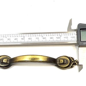 May include: An antique brass-colored metal drawer pull with a curved handle and decorative ends. A digital caliper is positioned over the handle, displaying a measurement of 4.8 inches on its screen, indicating the total length of the hardware.