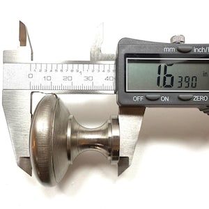 May include: A brushed nickel cabinet knob is measured with a digital caliper. The knob is approximately 1.6 inches in diameter. The caliper displays measurements in both inches and millimeters.