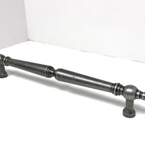 May include: A long, silver-toned metal cabinet pull handle with a weathered, antique finish. The handle features a central cylindrical bar with tapered ends, decorative ringed accents, and two rounded mounting posts at each end for attachment.