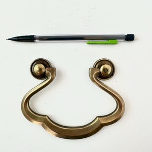 May include: A vintage-style brass drawer pull with a curved handle and two round mounting points. A black and clear mechanical pencil with a green grip is positioned above the drawer pull. The hardware is likely for furniture restoration.