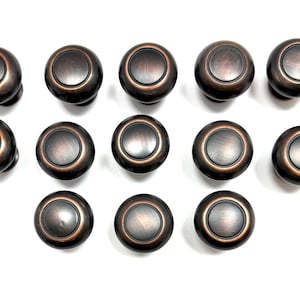 May include: A collection of fifteen bronze-colored cabinet knobs arranged on a white background. Each knob has a circular design with a slightly raised center and a dark, metallic finish. The knobs are evenly spaced.