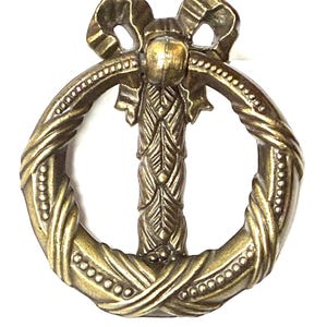 May include: A decorative brass-toned drawer pull with a bow-shaped top and a circular handle. The handle features a braided design with small beaded accents. The central support has a leaf-like pattern.