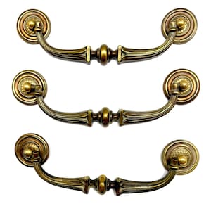 May include: Three antique brass drawer pulls with a decorative design. Each pull features a circular backplate with a detailed pattern, a curved handle, and a central decorative element. The hardware has a warm, aged finish, suggesting vintage or classic style.