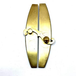May include: A vintage brass cabinet pull with an oval design. The pull is split into two halves, connected by a decorative curved handle with a small ball and circular accents. The brass has a warm, golden hue.