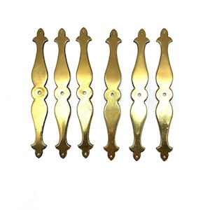 May include: Six vintage brass drawer pulls with a decorative design. Each pull is a warm gold color, featuring an elongated shape with a central circular detail and a decorative top and bottom. The pulls are arranged in a row against a white background.